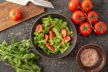 There is a bowl of light summer vegetable salad of tomatoes and greens and fresh ingredients for its cooking on a black background. Top view