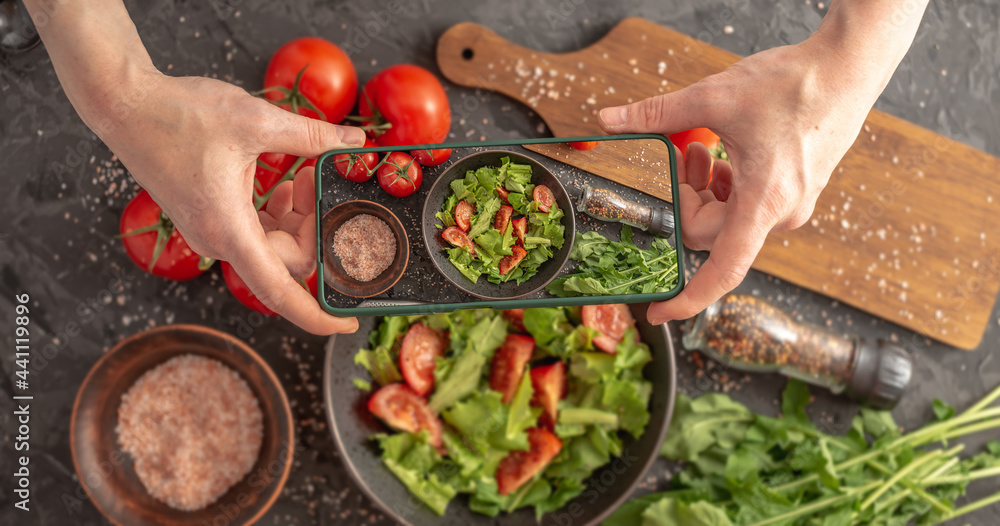 Wall mural women's hands are holding a mobile phone over a beautiful appetizing vegetable salad to take a photo