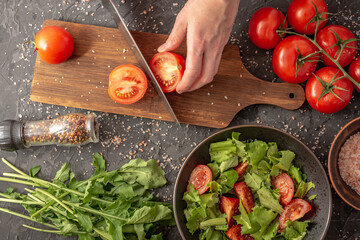 Women's hands are cutting a ripe tomato to cook a light healthy vegetable salad with tomatoes and...