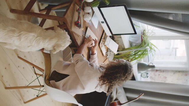 Vertical high angle shot of young woman working on computer at desk in home office and petting lovely golden retriever dog