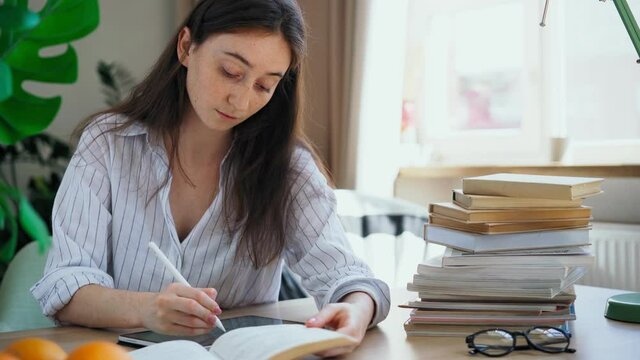 Young woman student preparing for exams rewrites a lecture from a book into a tablet with a stylus