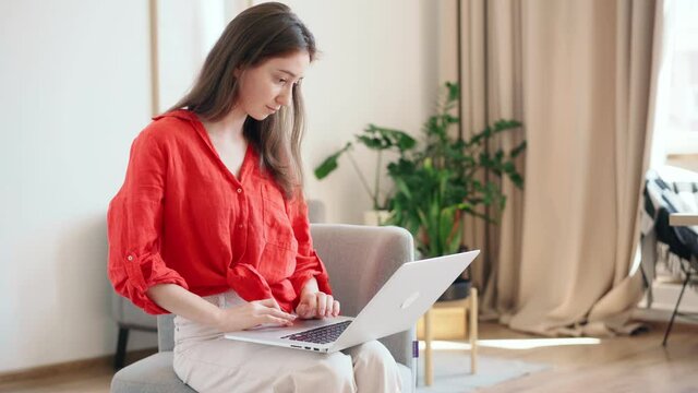 A beautiful young businesswoman in a bright red shirt is typing on a laptop while sitting in a comfortable armchair at the office