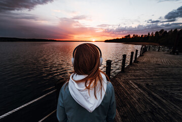 The girl listens to music with headphones stands on the pier near the lake and looks at the...