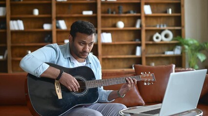 Smiling indian young man learning how to play guitar online, sitting on the sofa in front of the laptop, watching video tutorial, repeats exercises. E-studying and hobby online