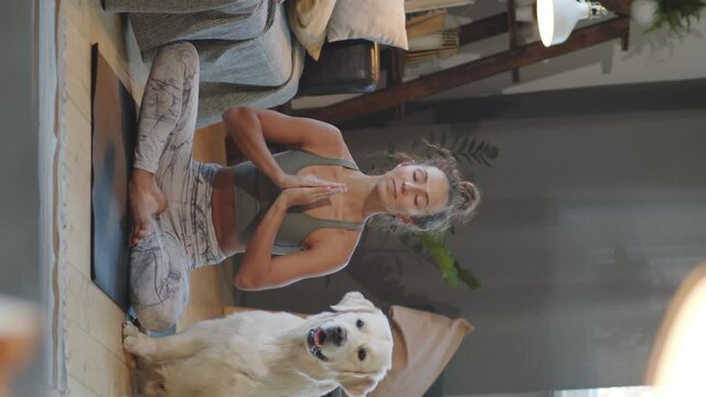 Vertical shot of young woman in sportswear sitting on mat near cute dog and practicing yoga meditation in lotus pose at home