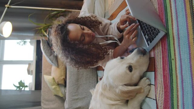 Vertical clip of young brunette woman lying on floor at home with lovely golden retriever dog and browsing the Internet on laptop