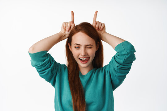 Close up portrait of sassy redhead girl shows bull horns gesture, winks and smiles daring, looking determined and confident, having fun, white background