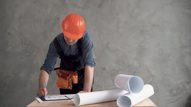 a male construction worker dressed in a uniform and an orange hard hat is bent over the project drawings and making adjustments with a pencil. Copy space.