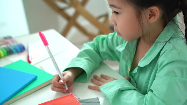 Asian girl sitting at table writing in school book at home