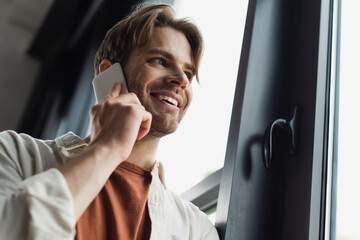 low angle view of smiling young man in beige shirt talking on cellphone near window in office