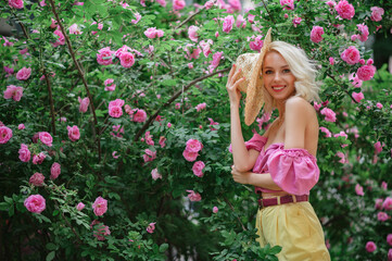Happy smiling beautiful blonde woman wearing pink top, holding straw hat, posing in blooming rose garden. Copy, empty space for text