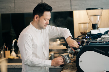 Professional barista holding metal jug warming milk using the coffee machine. Happy young man preparing coffee at counter.