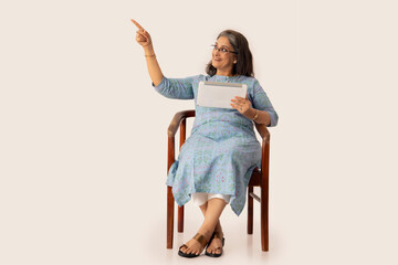 AN OLD WOMAN HAPPILY POINTING AWAY WHILE SITTING AND HOLDING DIGITAL TABLET