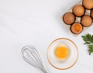 Banner. Eggs in a box, a glass cup with a raw egg and a whisk on a white background. Space for text, top view. The idea of ingredients for a recipe.