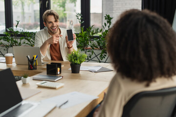 smiling young man pointing with finger to smartphone screen near blurred african american colleague in office