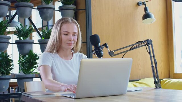 Front view of young cute woman with good mood communicating online in laptop with modern microphone in cafe. Concept of process working on radio with microphone and laptop. 
