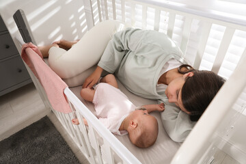 Tired young mother sleeping in crib with her baby at home, above view