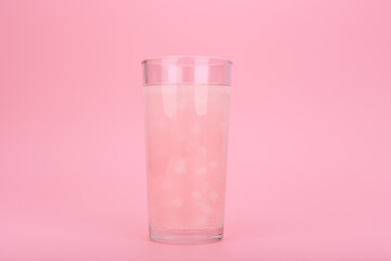 Close up of transparent glass with light pink fruit drink or lemonade with pieces of fruit against pink background. Summer composition. Concept of refreshing cold drinks for summer or detox drinks
