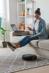 Robotic vacuum cleaner cleaning carpet, woman remote control mobile phone and enjoy rest, sitting...