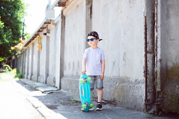 Stylish boy child in street clothes in a T-shirt, bandana, blue sunglasses and black and white sneakers with a turquoise skateboard in his hands against the background of a concrete wall.