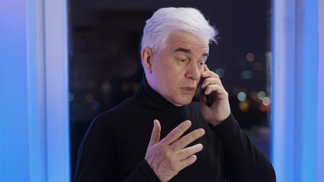 Serious old retired gentleman in turtleneck sweater speaks. Elder graying businessman by the window has business talk, holiday celebration fireworks. Grey hair male person. Senior man with white face.