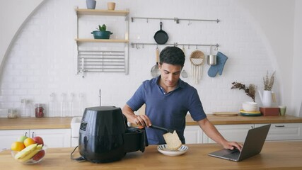 Handsome young caucasian man looking at instructions online on laptop on how to use modern air fryer to cook healthy food without using oil at home in kitchen