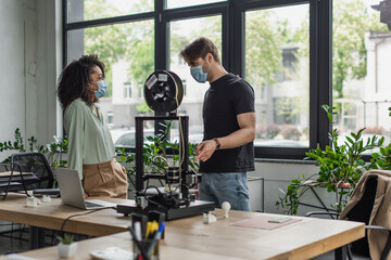 Fototapeta na wymiar interracial designers in medical masks talking near 3D printer in modern office
