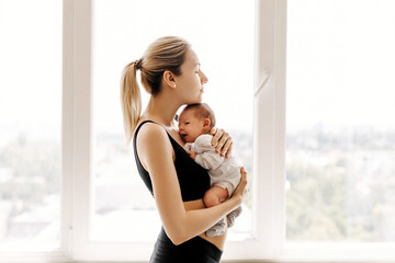Young mother holding and hugging newborn baby in her arms at home, by the window.