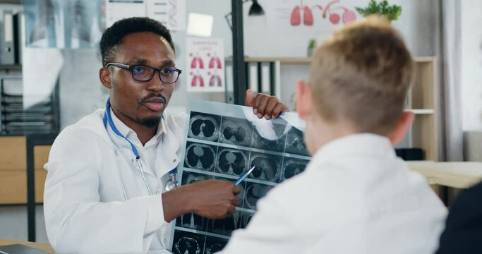 Handsome confident skillful black-skinned doctor in white gawn explaining diagnisis of his teen boy-patient with caring mother using boy's x-ray scan during their visit to medical office