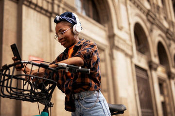 Happy young woman riding bicycle in the city. Beautiful african woman using the phone outdoors.