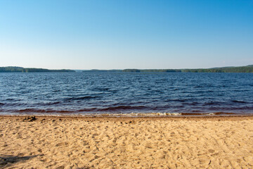 Varpaisniemi Beach's Sand, Water, and Sky. From the shore of Varpaisniemi Beach, you can see the brown sand, the wavy water, the green trees from the forest or islands and the clear blue sky.