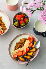 Delicious healthy breakfast, granola with yogurt and sweet fruits, banana and strawberries, blueberries and peaches, milk in a mug and flowers in a vase, top view