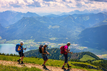 Unrecognizable people hiking on grass at Schafberg Railway with Lake Wolfgangsee and Dachstein Mountains against blue sky. Salzburg area, Austria.