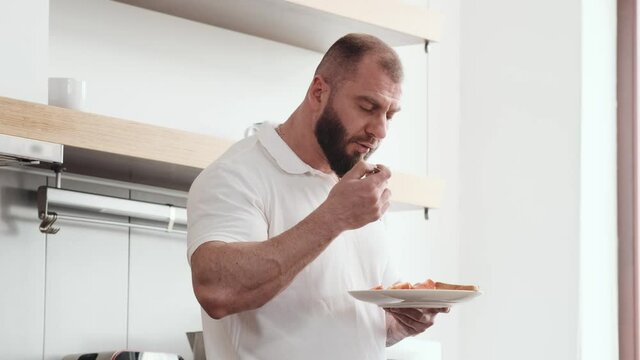 An attractive bearded man is eating breakfast standing in the kitchen at home
