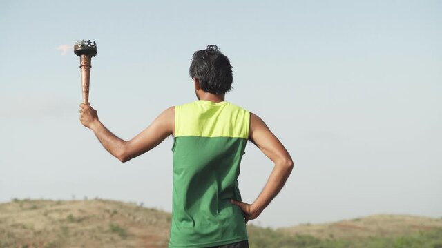 Back view of athlete proudly holding sports flame torch relay and looking mountains