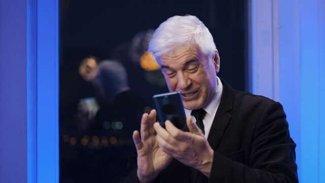 Smiling happy old retired gentleman in blazer suit looking at smartphone. Elder graying businessman by the window with fireworks. Grey haired male person. Senior gaffer man with white shaved face.