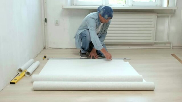 A man sitting on the floor cuts a roll of Wallpaper to paste Wallpaper on the wall during repairs in the apartment. Renovation of the apartment. interior design and Room renovations at house