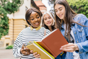 Obraz premium Enjoying university life. Positive friendly young African woman holding books and smiling while standing against university with her friends chatting about schedule classes.