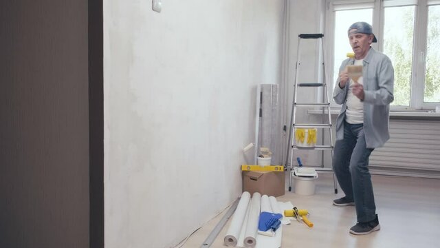 Positive man dancing and singing into a rubber roller having fun during home repair and painting. Construction site and renovation. House improvement and makeover concept. Room renovations at house