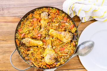 Aerial view of a rice with chicken and saffron or chicken paella in the classic paella pan and a ladle to serve on the white plate.