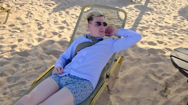Sweating is a concept. Funny businessman sniffs his armpit lying on a sun lounger on the beach in a shirt, tie, underwear and sunglasses