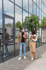 young interracial colleagues in medical masks walking with coffee paper cups near modern building