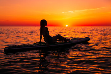 Girl on stand up paddle board at quiet sea with bright sunset or sunrise. Woman relaxing on sup...