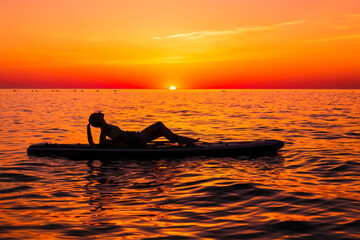 Girl on stand up paddle board at quiet sea with bright sunset or sunrise. Woman relaxing on sup board in sea.