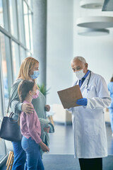 Mature pediatrician examining medical report while talking to mother and daughter at hospital hallway.