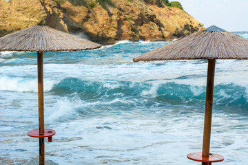 Two straw umbrellas on the beach near the stormy sea. European mountain coastline near the sea with...