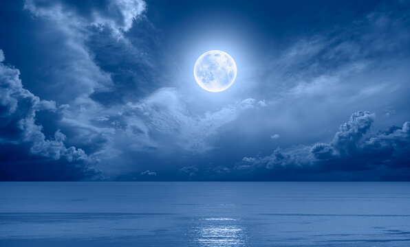 Night sky with full bright moon in the clouds, blue sea in the foreground "Elements of this image furnished by NASA"
