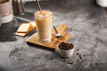 A glass with ice cubes and milk coffee on a wooden board, with brass straw and coffee beans