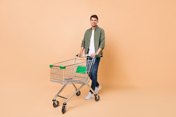 Full size photo of charming young happy man hold pushcart smile shopper isolated on beige color...