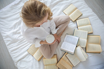 Beautiful sensual young woman with blonde hair wearing a white shirt reading books and drinking coffee on the bed in modern home